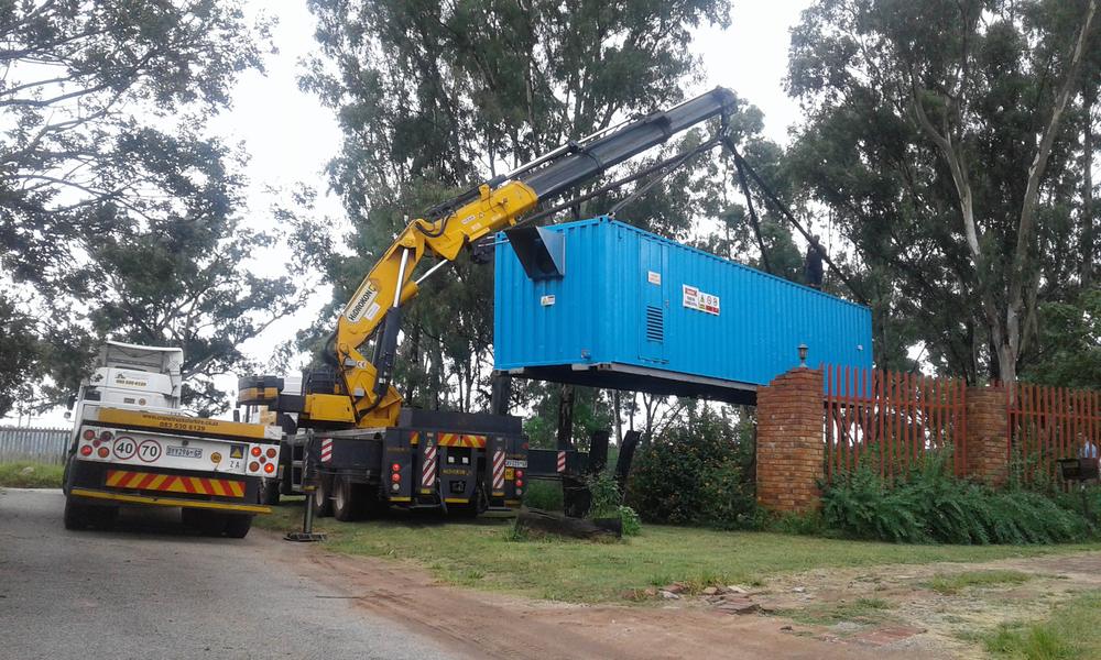 Yellow knuckle boom crane truck lifting a blue 12 metre container over a fence